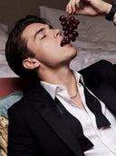 Valloria Kaptaan luxury perfume campaign featuring an elegant man in a black suit and unbuttoned shirt eating red grapes.
