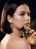 Elegant female model with a sleek black bob haircut holding a clear glass VALLORIA luxury perfume bottle against her shoulder in a dramatic, dark setting.